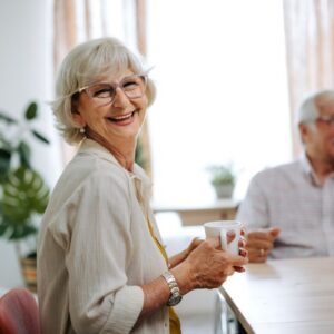An older woman smiles at the camera as she enjoys coffee with other senior living residents.